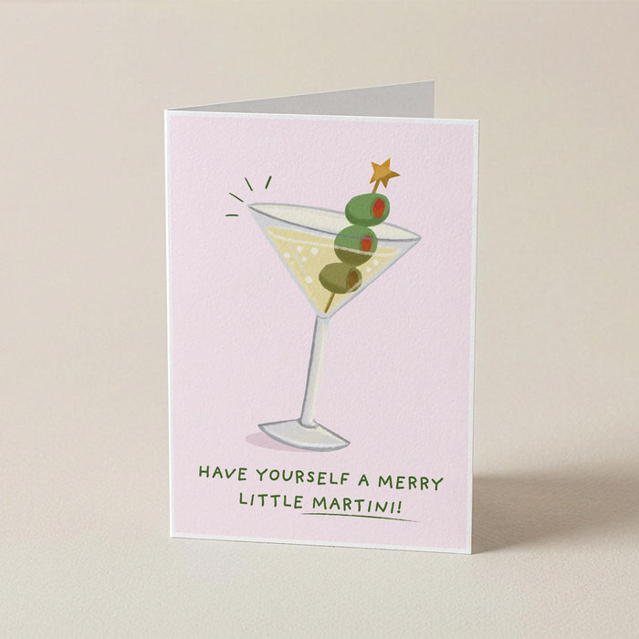 A folded greeting card standing upright on a light beige surface. The card has a pale pink background and features an illustration of a martini glass with green olives and a small gold star, with the text 'HAVE YOURSELF A MERRY LITTLE MARTINI!!' printed below.
