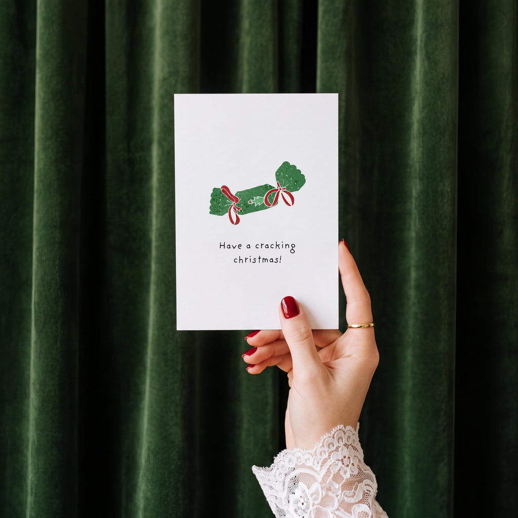A hand with red nail polish and a white lace cuff holds up a white Christmas greeting card that says 'Have a cracking Christmas!' with a green ornament illustration, set against a dark green velvet curtain background.