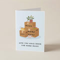 A folded greeting card standing upright on a beige surface. The card features an illustration of stacked cardboard boxes with the words 'MUGS' on them, a small potted plant on top, and the text 'NOW YOU HAVE ROOM FOR MORE MUGS!' below.