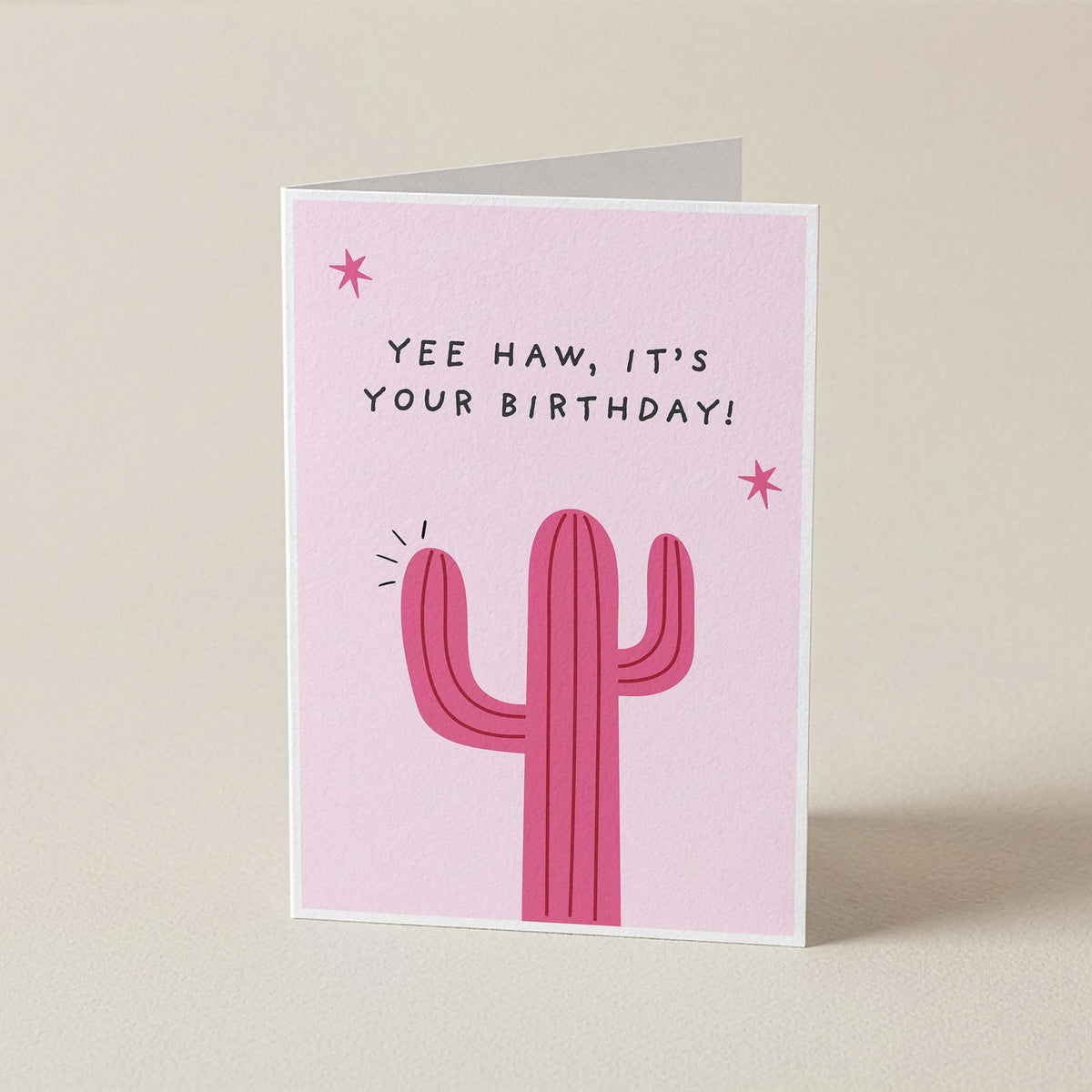 A pink greeting card with a cactus illustration and the text 'YEE HAW, IT'S YOUR BIRTHDAY!' standing upright on a light beige background.