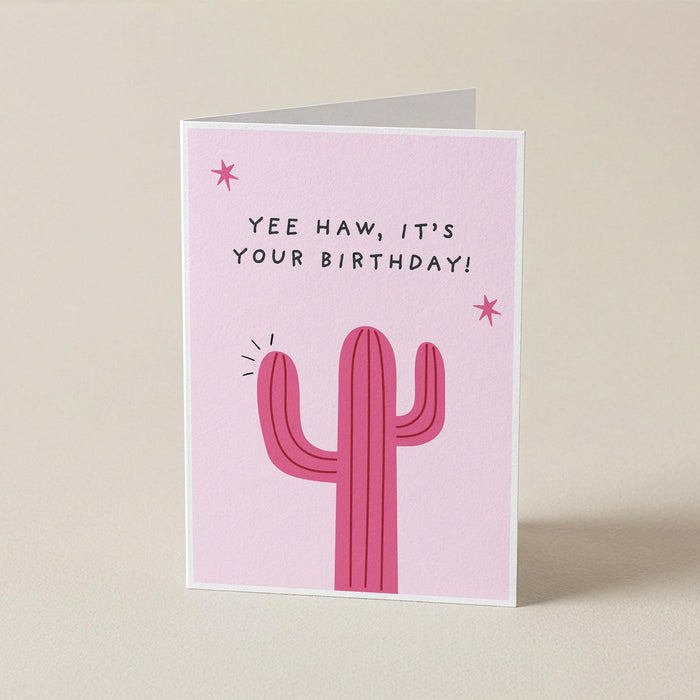 A pink greeting card with a cactus illustration and the text 'YEE HAW, IT'S YOUR BIRTHDAY!' standing upright on a light beige background.