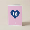 A folded greeting card standing upright on a light beige surface. The card has a pink front with a blue heart-shaped balloon graphic containing the white number 18.