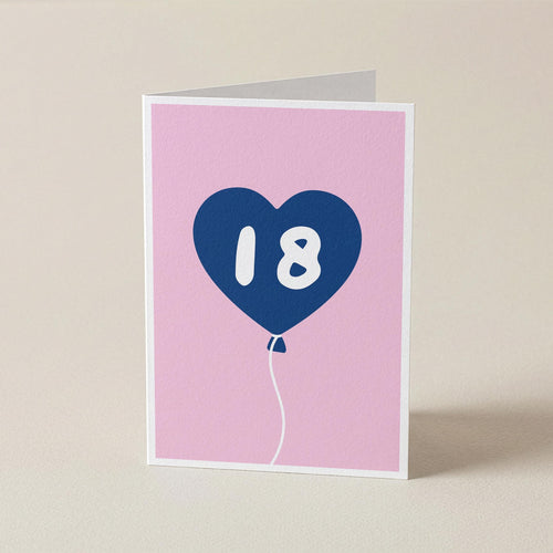 A folded greeting card standing upright on a light beige surface. The card has a pink front with a blue heart-shaped balloon graphic containing the white number 18.