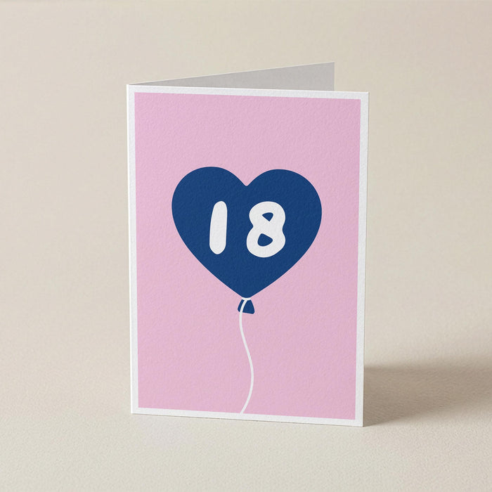 A folded greeting card standing upright on a light beige surface. The card has a pink front with a blue heart-shaped balloon graphic containing the white number 18.
