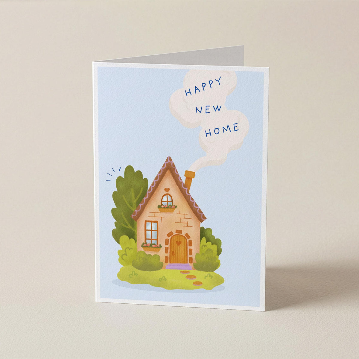 A folded greeting card standing upright on a light beige surface. The card features a cute illustration of a small house with green bushes and a white speech bubble that reads 'HAPPY NEW HOME'.