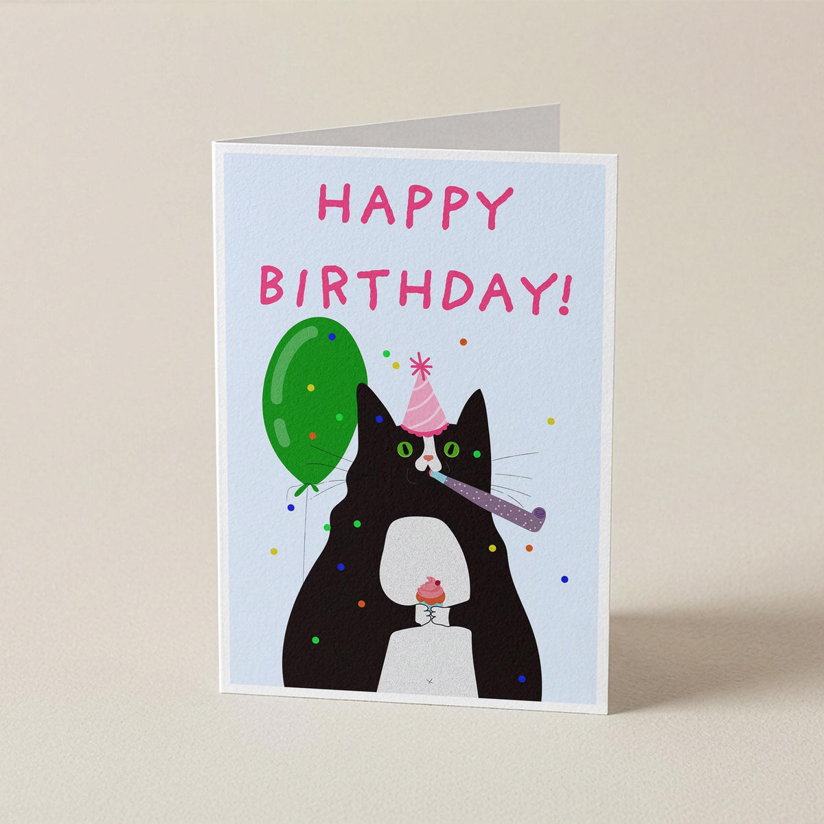 A folded greeting card standing upright on a light beige surface. The card features a black cat wearing a pink party hat and blowing a purple party horn, with a green balloon and colorful confetti, and the words 'HAPPY BIRTHDAY!' in pink at the top.
