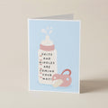 A folded greeting card standing upright on a light beige surface. The card has a pale blue front with a simple illustration of a white baby bottle with pink details and a pink pacifier, plus the text 'SHITS AND GIGGLES ARE COMING YOUR WAY!'.