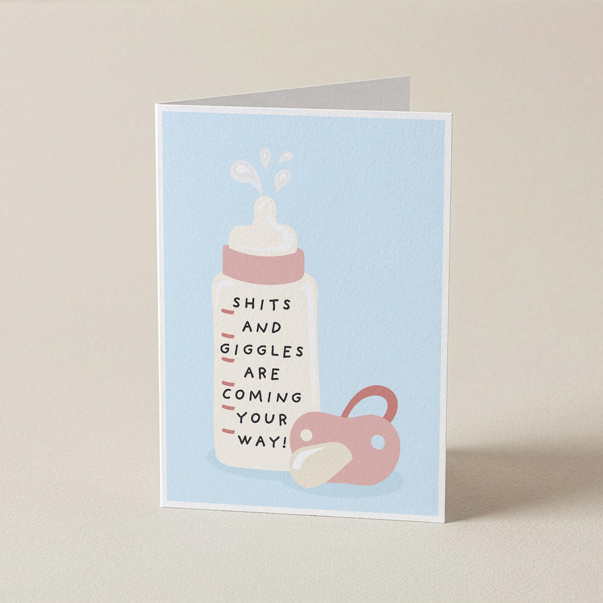 A folded greeting card standing upright on a light beige surface. The card has a pale blue front with a simple illustration of a white baby bottle with pink details and a pink pacifier, plus the text 'SHITS AND GIGGLES ARE COMING YOUR WAY!'.