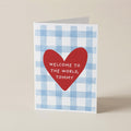 A folded greeting card standing upright on a light beige surface. The card has a white and light blue gingham pattern, with a large red heart in the center containing the white text 'WELCOME TO THE WORLD, TOMMY'.