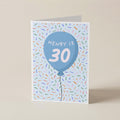 A folded greeting card standing upright on a beige surface. The card features a light blue balloon graphic with the text 'HENRY IS 30' in white, surrounded by multicolored confetti on a pale blue background.