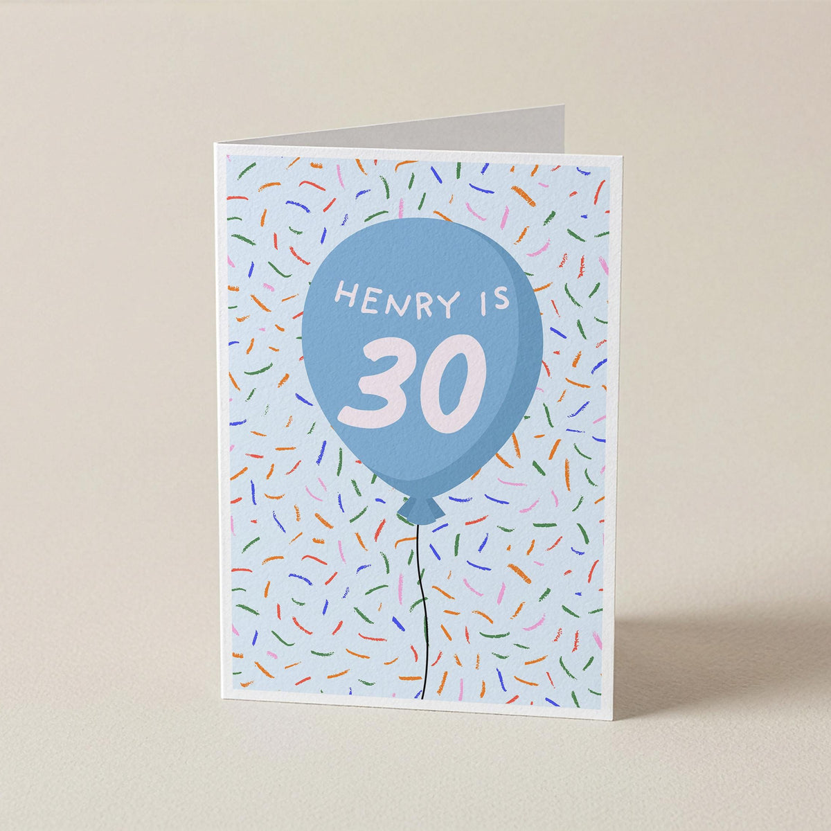 A folded greeting card standing upright on a beige surface. The card features a light blue balloon graphic with the text 'HENRY IS 30' in white, surrounded by multicolored confetti on a pale blue background.