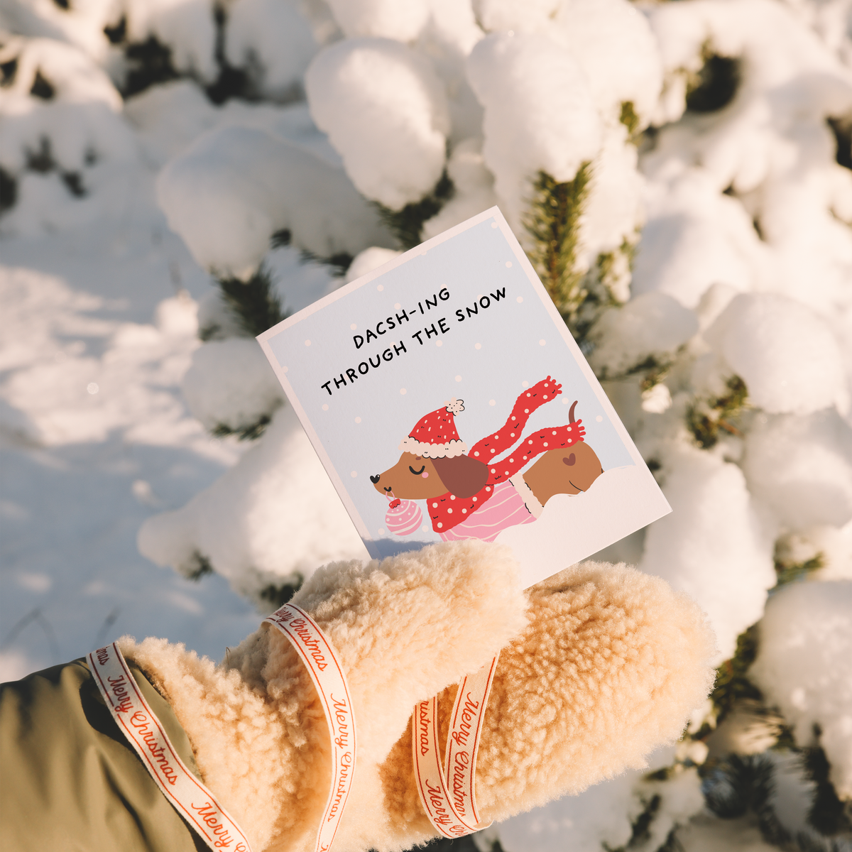 A hand wearing cream-colored fleece mittens with red and white striped ribbons holds a square greeting card that reads 'DACHSHING THROUGH THE SNOW' above an illustrated dachsh dog in a red Santa hat and scarf, set against a snowy outdoor background.