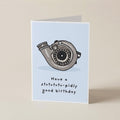 A folded greeting card standing upright on a light beige surface. The card has a pale blue front with a gray turbocharger illustration and the text 'Have a stutututu-pidly good birthday'.
