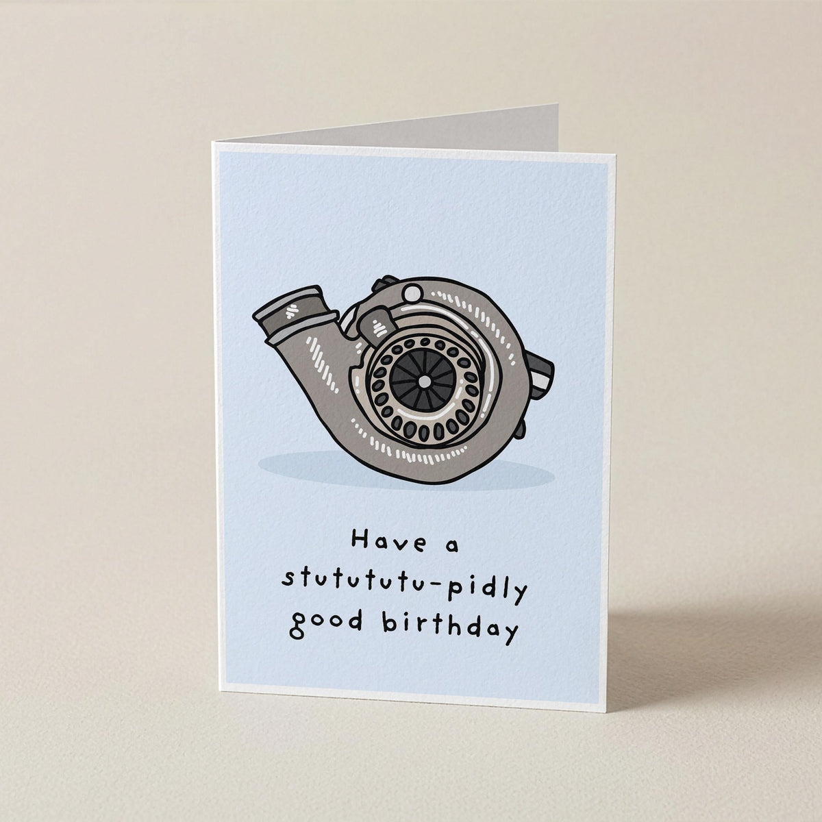A folded greeting card standing upright on a light beige surface. The card has a pale blue front with a gray turbocharger illustration and the text 'Have a stutututu-pidly good birthday'.