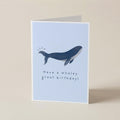 A folded greeting card standing upright on a light beige surface. The card has a pale blue front with a dark blue whale illustration and the text 'Have a whaley great birthday!'.