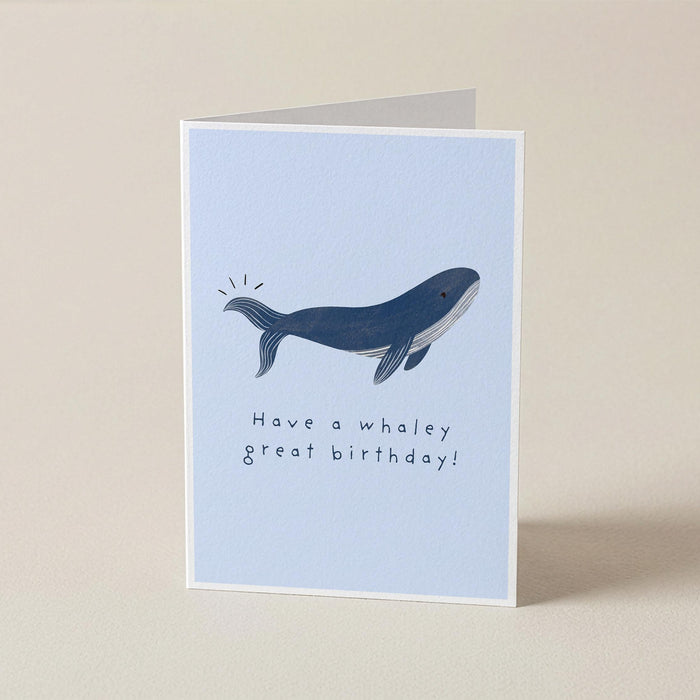 A folded greeting card standing upright on a light beige surface. The card has a pale blue front with a dark blue whale illustration and the text 'Have a whaley great birthday!'.