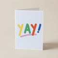 A white greeting card standing upright on a beige surface, featuring the word 'YAY!' in colorful playful letters with a pink underline and blue exclamation mark.