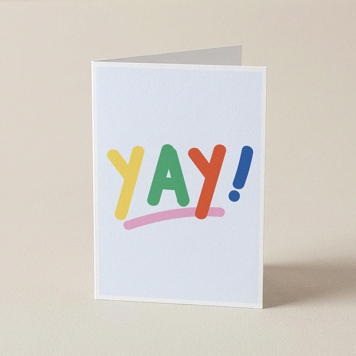A white greeting card standing upright on a beige surface, featuring the word 'YAY!' in colorful playful letters with a pink underline and blue exclamation mark.
