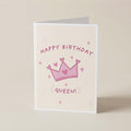 A folded greeting card standing upright on a light beige surface. The card has a cream background with a pink crown illustration, small pink stars, and the words 'HAPPY BIRTHDAY QUEEN!' in pink lettering.