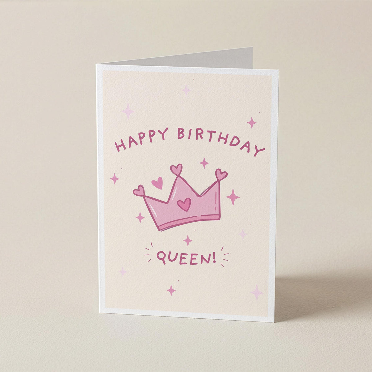 A folded greeting card standing upright on a light beige surface. The card has a cream background with a pink crown illustration, small pink stars, and the words 'HAPPY BIRTHDAY QUEEN!' in pink lettering.