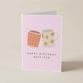 A pink greeting card standing upright on a light beige surface. The card features an illustration of two mugs, one orange-brown with a grid pattern and one cream with yellow flowers, above the text 'HAPPY BIRTHDAY BEST TEA'.
