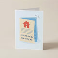 A folded greeting card standing upright on a light beige surface. The card features a minimalist illustration of a mortgage document with a red house icon and the text 'MORTGAGE WANKERS' in dark blue lettering.