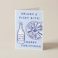 A folded Christmas greeting card standing upright on a light beige surface. The card has a pale blue background with dark blue line drawings of a bottle and a plate of nuts, with the text 'DRINKS & PICKY BITS! MERRY CHRISTMAS!'.