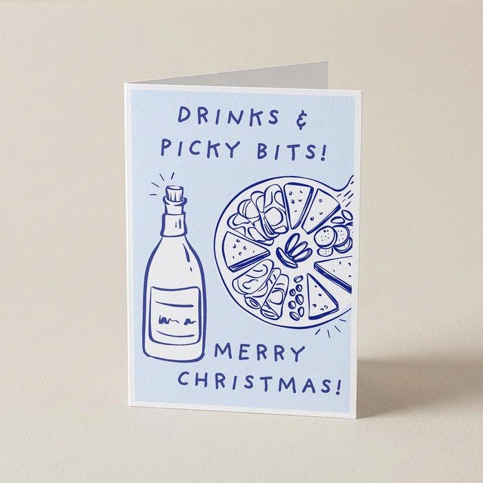 A folded Christmas greeting card standing upright on a light beige surface. The card has a pale blue background with dark blue line drawings of a bottle and a plate of nuts, with the text 'DRINKS & PICKY BITS! MERRY CHRISTMAS!'.
