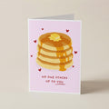 A folded greeting card standing upright on a light beige surface. The card has a pale pink background with an illustration of a stack of pancakes topped with butter and syrup, small red hearts, and the text 'NO ONE STACKS UP TO YOU' near the bottom.
