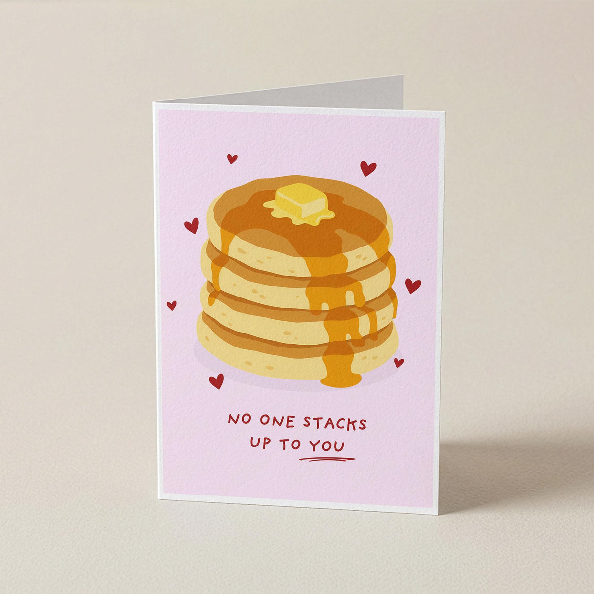 A folded greeting card standing upright on a light beige surface. The card has a pale pink background with an illustration of a stack of pancakes topped with butter and syrup, small red hearts, and the text 'NO ONE STACKS UP TO YOU' near the bottom.