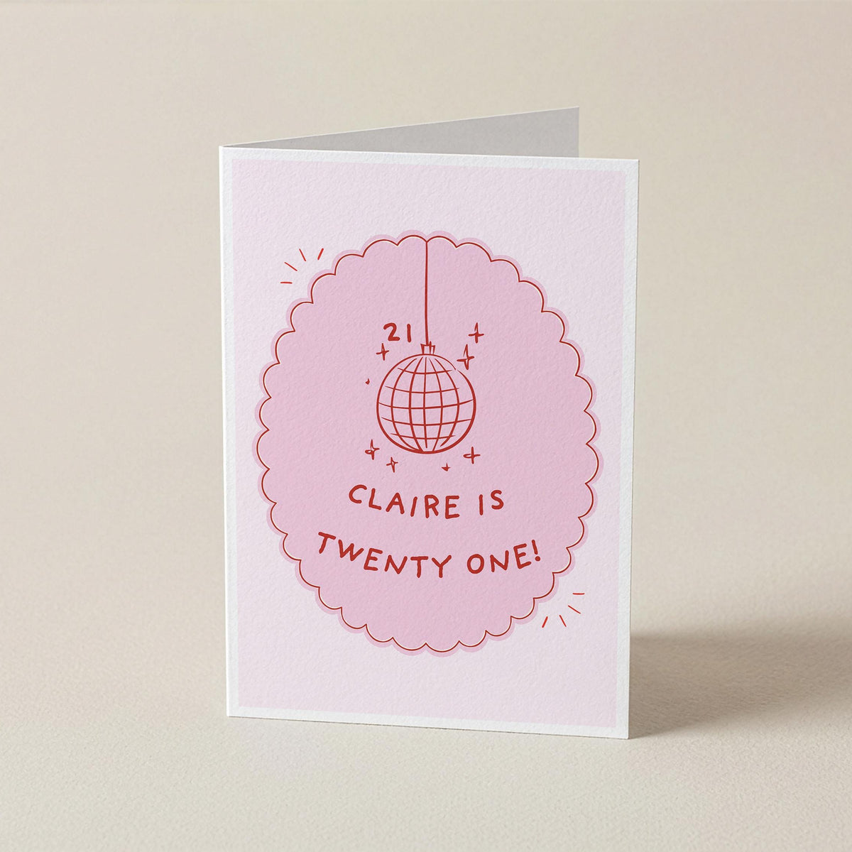 A folded greeting card standing upright on a light beige surface. The card is white with a pink scalloped oval graphic featuring a disco ball illustration and the text 'CLAIRE IS TWENTY ONE!'.