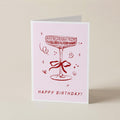 A folded birthday card standing upright on a light beige surface. The card has a pale pink background with a red line drawing of a cocktail glass tied with a bow and the words 'HAPPY BIRTHDAY!' printed at the bottom.