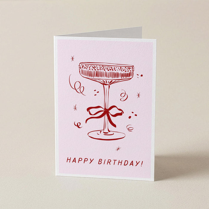 A folded birthday card standing upright on a light beige surface. The card has a pale pink background with a red line drawing of a cocktail glass tied with a bow and the words 'HAPPY BIRTHDAY!' printed at the bottom.