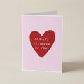A folded greeting card standing upright on a light beige surface. The card front is pale pink with a large red heart in the center and white text reading 'ALWAYS BELIEVED IN YOU.