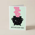 A folded greeting card standing upright on a light beige surface. The card features a black cauldron with white stars and a pink smoking shape above it, with the text 'WITCHES DON'T AGE.