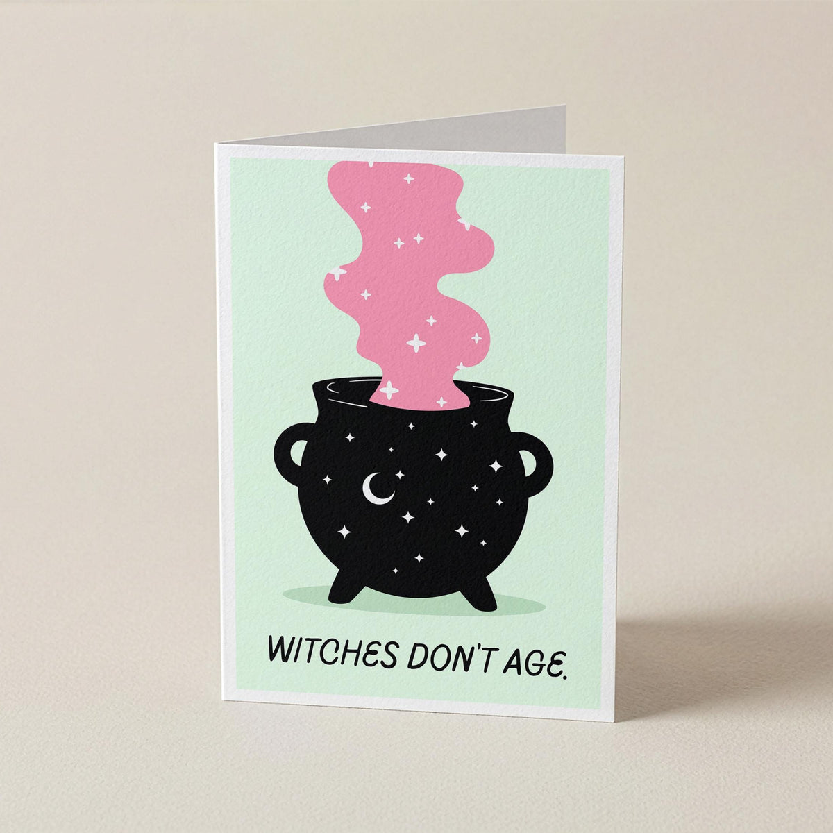 A folded greeting card standing upright on a light beige surface. The card features a black cauldron with white stars and a pink smoking shape above it, with the text 'WITCHES DON'T AGE.