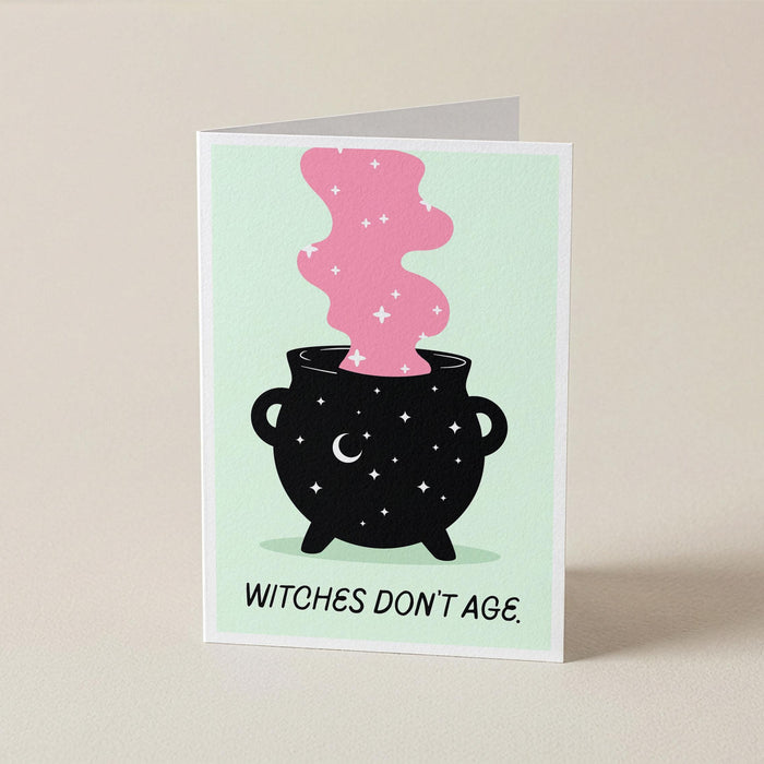 A folded greeting card standing upright on a light beige surface. The card features a black cauldron with white stars and a pink smoking shape above it, with the text 'WITCHES DON'T AGE.