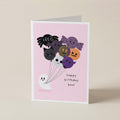 A folded greeting card standing upright on a light beige surface. The card has a pink front with a cute illustration of Halloween-themed balloons and a small white ghost, plus the text 'happy birthday boo!'.