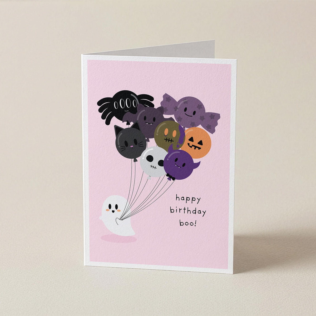 A folded greeting card standing upright on a light beige surface. The card has a pink front with a cute illustration of Halloween-themed balloons and a small white ghost, plus the text 'happy birthday boo!'.