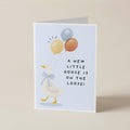 A folded greeting card standing upright on a light beige surface. The card features a white goose wearing a blue bow tie and holding three balloons in yellow, orange, and blue, with the text 'A NEW LITTLE GOOSE IS ON THE LOOSE!' printed in black.