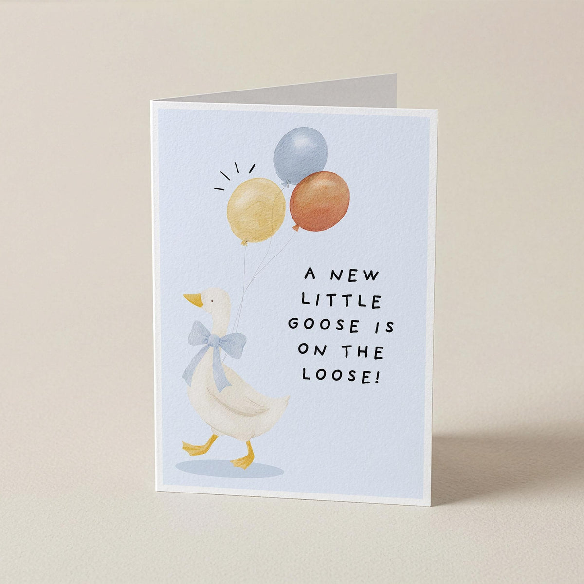 A folded greeting card standing upright on a light beige surface. The card features a white goose wearing a blue bow tie and holding three balloons in yellow, orange, and blue, with the text 'A NEW LITTLE GOOSE IS ON THE LOOSE!' printed in black.