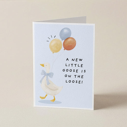A folded greeting card standing upright on a light beige surface. The card features a white goose wearing a blue bow tie and holding three balloons in yellow, orange, and blue, with the text 'A NEW LITTLE GOOSE IS ON THE LOOSE!' printed in black.