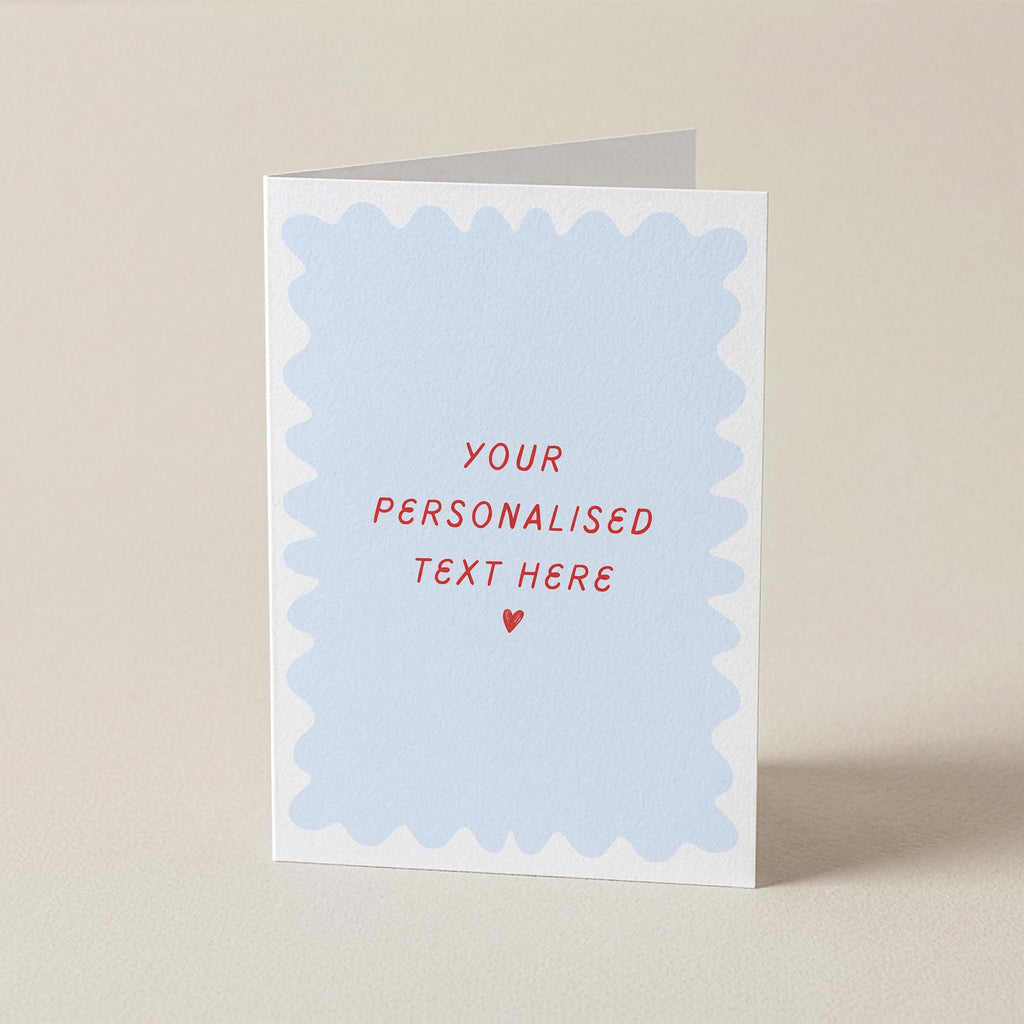 A folded greeting card standing upright on a light beige surface. The front has a white border with a pale blue scalloped inner panel and red text reading 'YOUR PERSONALISED TEXT HERE' with a small red heart below.