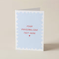A folded greeting card standing upright on a light beige surface. The front has a white border with a pale blue scalloped inner panel and red text reading 'YOUR PERSONALISED TEXT HERE' with a small red heart below.