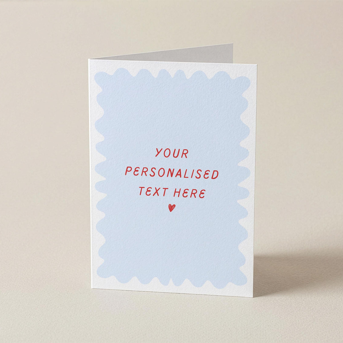 A folded greeting card standing upright on a light beige surface. The front has a white border with a pale blue scalloped inner panel and red text reading 'YOUR PERSONALISED TEXT HERE' with a small red heart below.