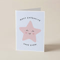 A folded greeting card standing upright on a light beige surface. The card has a white border and features a pink star illustration with a smiling face, with the text 'MOST EXPENSIVE' above and 'SHAG EVER!' below.