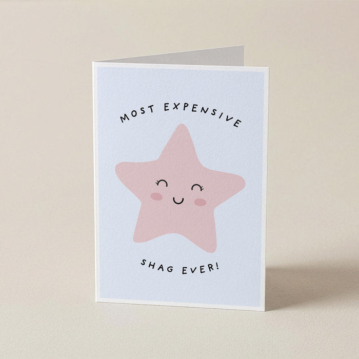 A folded greeting card standing upright on a light beige surface. The card has a white border and features a pink star illustration with a smiling face, with the text 'MOST EXPENSIVE' above and 'SHAG EVER!' below.