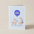 A folded greeting card standing upright on a light beige surface. The card features a light blue illustration with two cartoon chickens wearing party hats and holding a blue 'Happy Birthday' balloon, with one chicken also holding a small birthday cake.