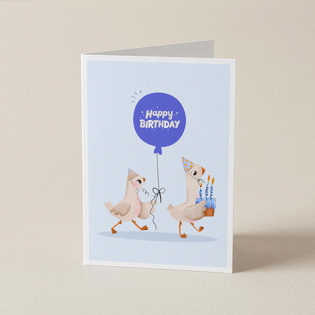 A folded greeting card standing upright on a light beige surface. The card features a light blue illustration with two cartoon chickens wearing party hats and holding a blue 'Happy Birthday' balloon, with one chicken also holding a small birthday cake.