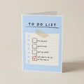 A folded greeting card standing upright on a light beige surface. The card has a pale blue border and reads 'TO DO LIST' above a checklist with a tape graphic and a red checkmark on the last item.